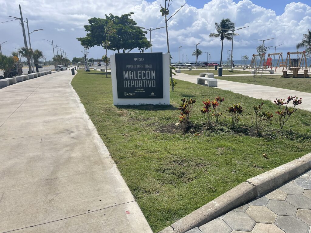 Paseo peatonal y áreas verdes del Malecón Deportivo de Santo Domingo, con señalización, palmeras y el mar al fondo.