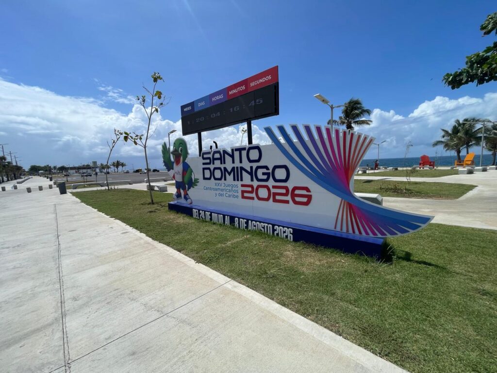 Juegos Centroamericanos y del Caribe Santo Domingo 2026: calendario, sedes, entradas y cómo asistir Valla con cuenta regresiva y el logotipo de los XXV Juegos Centroamericanos y del Caribe Santo Domingo 2026, instalada al aire libre junto a un paseo costero, con palmeras, mobiliario urbano y el mar al fondo bajo un cielo despejado.